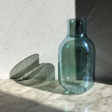 Jarrón con forma de botella de 24 cm de altura y 12cm de diámetro, de color verde azulado y con reflejos iridiscentes, fotografiado sobre un mármol y a la luz del sol